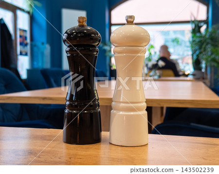 Black and white pepper grinders stand on a wooden table inside a cozy restaurant, highlighting a minimalist culinary accessory set for seasoning meals in a professional dining room Black and white pepper grinders stand on a wooden table inside a cozy restaurant, highlighting a minimalist culinary accessory set for seasoning meals in a professional dining room 143502239