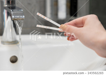 A hand holds testing strips near a running bathroom faucet, measuring water quality and mineral content for home health safety assessment in a clean, modern residential environment A hand holds testing strips near a running bathroom faucet, measuring water quality and mineral content for home health safety assessment in a clean, modern residential environment 143502283