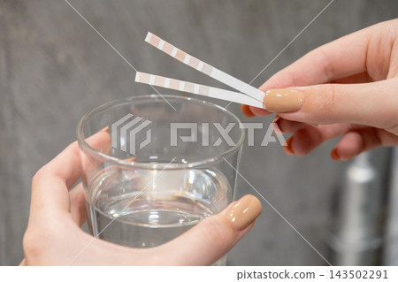 A pair of hands holding chemical test strips above a glass of clear water to check quality and safety levels in a laboratory or home environment during a routine hygiene evaluation A pair of hands holding chemical test strips above a glass of clear water to check quality and safety levels in a laboratory or home environment during a routine hygiene evaluation 143502291