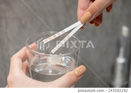 A hand holds a test strip over a glass of clear liquid to check water quality, representing a simple home health assessment procedure for safety analysis in a clean bathroom setting A hand holds a test strip over a glass of clear liquid to check water quality, representing a simple home health assessment procedure for safety analysis in a clean bathroom setting 143502292