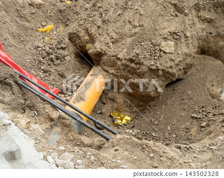 Orange sewer pipe and black cables exposed in a deep ground trench at a road construction site, representing utility infrastructure maintenance work during urban development project Orange sewer pipe and black cables exposed in a deep ground trench at a road construction site, representing utility infrastructure maintenance work during urban development project 143502328