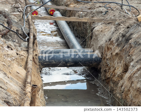 Industrial water pipes installed in a deep muddy construction trench for urban infrastructure repair, providing a clear view of heavy utility maintenance work during a site development Industrial water pipes installed in a deep muddy construction trench for urban infrastructure repair, providing a clear view of heavy utility maintenance work during a site development 143502373
