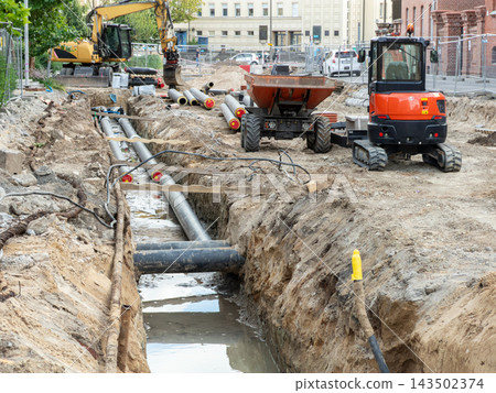 A construction site featuring an open trench with large underground pipes and heavy machinery, representing urban infrastructure repair and utility renovation work in a city environment A construction site featuring an open trench with large underground pipes and heavy machinery, representing urban infrastructure repair and utility renovation work in a city environment 143502374