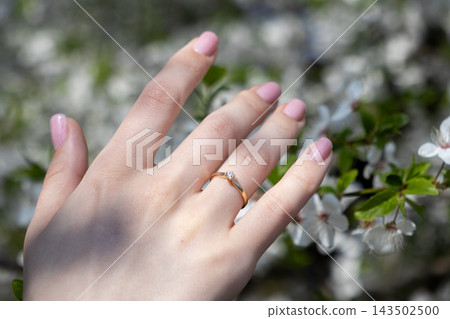 A gold diamond engagement ring on a person finger with light pink manicure, set against a soft blurred background of white blooming spring cherry tree flowers, symbolizing romance A gold diamond engagement ring on a person finger with light pink manicure, set against a soft blurred background of white blooming spring cherry tree flowers, symbolizing romance 143502500