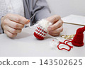 Hands of a person sewing together knitted parts of a Tomte doll made from yarn. 143502625