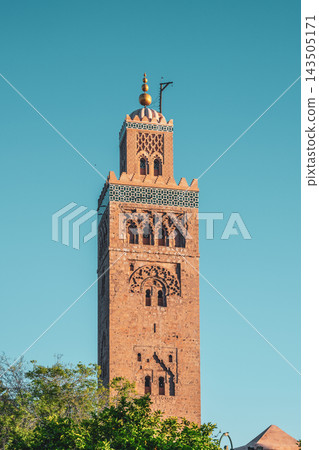 Kutubiyya Mosque stands tall in Marrakesh. The sun shines on its famous tower. The surrounding trees add green to the scene as visitors explore the area. Kutubiyya Mosque stands tall in Marrakesh. The sun shines on its famous tower. The surrounding trees add green to the scene as visitors explore the area. 143505171