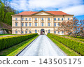 Boskovice Castle stands in Czechia with its classic Empire building style. The pathway leads to the front entrance, surrounded by trees and green bushes on a clear day. 143505175