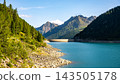 Lake Neves sits at the end of the Molini Valley in the Zillertal Alps, Italy. It features clear blue water and surrounding mountains with green trees. People may walk along the shore. 143505178