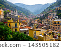 People enjoy the view of Riomaggiore village in Cinque Terre, Italy. The colorful buildings line the streets, surrounded by mountains and greenery. It is a bright day. 143505182