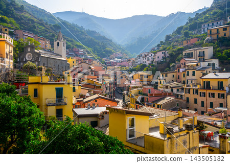 People enjoy the view of Riomaggiore village in Cinque Terre, Italy. The colorful buildings line the streets, surrounded by mountains and greenery. It is a bright day. People enjoy the view of Riomaggiore village in Cinque Terre, Italy. The colorful buildings line the streets, surrounded by mountains and greenery. It is a bright day. 143505182