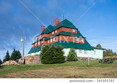 This is Masaryk's cottage located on Serlich. It is a well-known mountain cottage in the Orlicke ountains. The structure features multiple roofs and is surrounded by grass and trees. 143505187