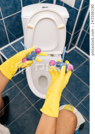 Vertical shot of hands in protective gloves inserting blue and pink toilet freshener block into toilet bowl, representing essential household cleaning, hygiene maintenance, routine bathroom care. Vertical shot of hands in protective gloves inserting blue and pink toilet freshener block into toilet bowl, representing essential household cleaning, hygiene maintenance, routine bathroom care. 143506190
