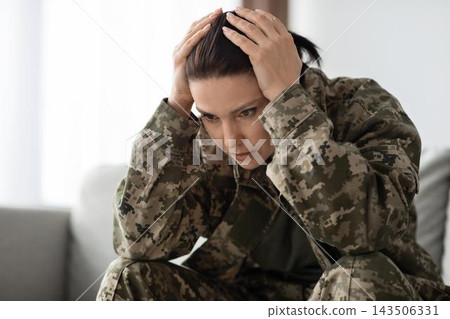 Worried Female Soldier In Military Uniform Showing Distress By Placing Hands On Head While Sitting On Couch In Living Room, Illustrating Mental Health Challenges Faced By Service Members. 143506331