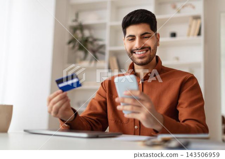 Happy Young Man With Dark Hair And Beard Using Smartphone While Holding Credit Card At His Workplace In Modern Office, Engaged In Online Shopping Or Banking, Business Concept. Happy Young Man With Dark Hair And Beard Using Smartphone While Holding Credit Card At His Workplace In Modern Office, Engaged In Online Shopping Or Banking, Business Concept. 143506959