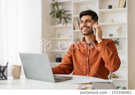 Smiling Young Man Of Middle Eastern Descent Talking On Phone While Sitting At Desk With Laptop, Engaged In Professional Work At Stylish Office, Reflecting Positive Business Concept. Smiling Young Man Of Middle Eastern Descent Talking On Phone While Sitting At Desk With Laptop, Engaged In Professional Work At Stylish Office, Reflecting Positive Business Concept. 143506961