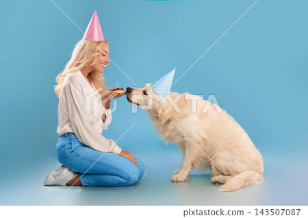 Smiling Young Caucasian Woman Joyfully Feeding Birthday Cake To Her Golden Retriever In Bright Studio Setting, Celebrating Pet's Special Day With Happiness And Love. Celebration Concept. 143507087