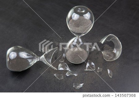 Broken hourglasses with dark sand and shattered glass scattered on a dark surface Broken hourglasses with dark sand and shattered glass scattered on a dark surface 143507308