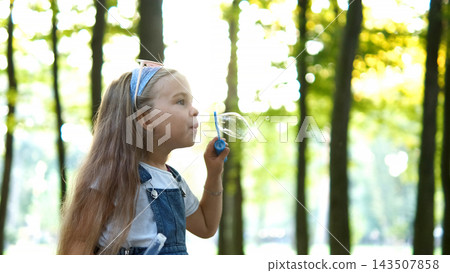 Little happy child girl blowing soap bubbles outside in green park. Outdoor summer activities concept. Little happy child girl blowing soap bubbles outside in green park. Outdoor summer activities concept. 143507858