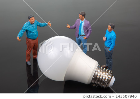 Miniature people gather around a large light bulb in a casual discussion setting Miniature people gather around a large light bulb in a casual discussion setting 143507940