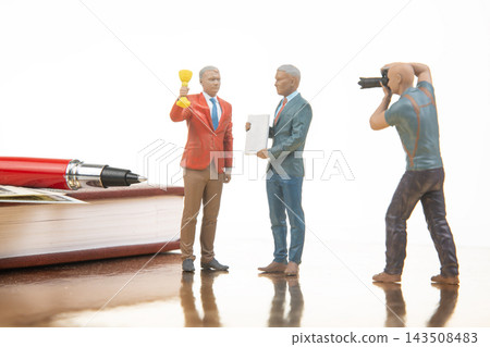 Miniature figures celebrate an achievement with a trophy and a camera in a simple office setting Miniature figures celebrate an achievement with a trophy and a camera in a simple office setting 143508483