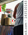 The backs of street food vendors preparing meals at a bustling outdoor stall, with stacks of dishes piled up. 143510143