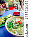 A steaming bowl of Vietnamese pho, served on a blue plastic table, garnished with chili peppers and fresh herbs. 143510144