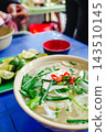 Delicious Vietnamese pho on a blue table, garnished with green herbs and vibrant red chilies. 143510145
