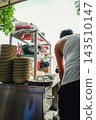 A wide-angle shot capturing the lively atmosphere, showing street food vendors working at a bustling stall with stacks of dishes. 143510147