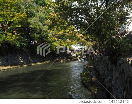 A tranquil waterside landscape viewed from the promenade along the Hachiman Canal in Omihachiman, featuring imposing stone walls and a tunnel of greenery. A tranquil waterside landscape viewed from the promenade along the Hachiman Canal in Omihachiman, featuring imposing stone walls and a tunnel of greenery. 143512142