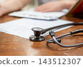A black stethoscope metal chestpiece rests medical paperwork wooden desk while person writes the softly blurred background suggesting clinical records consultation 143512307