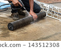 A person crouches beside cylindrical air conditioner blower uses spray nozzle to wash its dark fins water wet concrete floor other white components resting nearby 143512308
