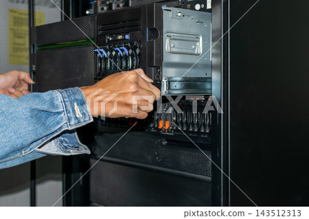 A technician wearing denim sleeve works both hands inside an open server rack stacked drive bays metal panels cables visible close view of computer equipment maintenance A technician wearing denim sleeve works both hands inside an open server rack stacked drive bays metal panels cables visible close view of computer equipment maintenance 143512313