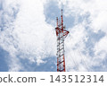 A red white lattice communications tower rises into blue sky scattered white clouds while antenna equipment cross braces reveal the structure of the telecommunications mast 143512314