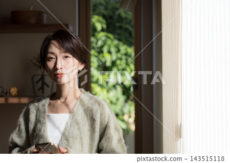 一幅日本女性的肖像畫,她站在自家窗邊,沐浴在自然光中,身穿秋冬針織衫或開襟衫。 一幅日本女性的肖像畫,她站在自家窗邊,沐浴在自然光中,身穿秋冬針織衫或開襟衫。 143515118