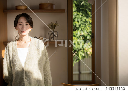 A portrait of a Japanese woman standing by a window in her home, bathed in natural light, wearing an autumn/winter knit sweater and looking directly at the camera. A portrait of a Japanese woman standing by a window in her home, bathed in natural light, wearing an autumn/winter knit sweater and looking directly at the camera. 143515130