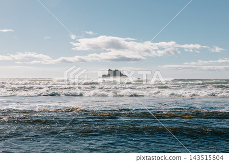 Ocean waves crashing near sea stack rock formation under cloudy blue sky 143515804