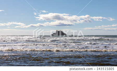 Ocean waves crashing near a rocky sea stack offshore under a cloudy blue sky Ocean waves crashing near a rocky sea stack offshore under a cloudy blue sky 143515805