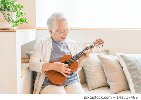 A senior woman with gray hair enjoys playing the ukulele in her living room at home. A life filled with hobbies and music. A senior woman with gray hair enjoys playing the ukulele in her living room at home. A life filled with hobbies and music. 143517469