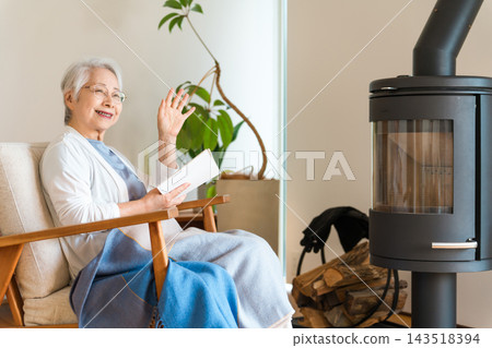 一位頭髮花白的老婦人面帶微笑,一邊揮手致意,一邊在客廳裡捧著一本書,客廳裡燃著柴火爐。多麼溫暖的冬日生活啊! 一位頭髮花白的老婦人面帶微笑,一邊揮手致意,一邊在客廳裡捧著一本書,客廳裡燃著柴火爐。多麼溫暖的冬日生活啊! 143518394