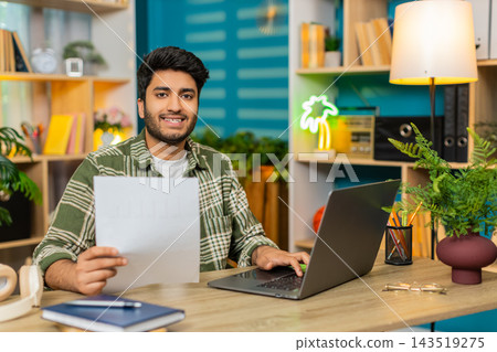 Indian man on home table typing on laptop holding document and smiling at camera in portrait view Indian man on home table typing on laptop holding document and smiling at camera in portrait view 143519275
