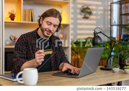 Young man on home table entering card data on laptop for online shopping with happy satisfied smile Young man on home table entering card data on laptop for online shopping with happy satisfied smile 143519277