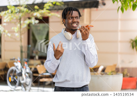 African American young man talking on smartphone speaker walking in city street with hand gestures African American young man talking on smartphone speaker walking in city street with hand gestures 143519534