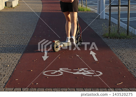 Electric scooter traveling along a dedicated cycling path marked with bicycle symbols and directional arrows, urban micromobility, road safety, shared routes, and modern city infrastructure. 143520275