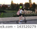 Woman wearing a helmet rides an electric scooter along a paved urban path at sunset, carrying a pink backpack beside landscaped greenery, trees and tall ornamental grasses. 143520279