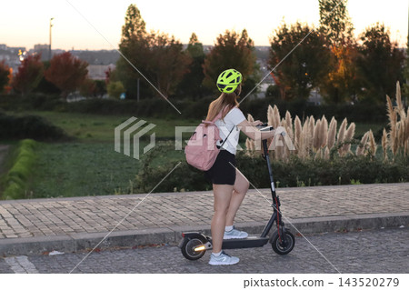 Woman wearing a helmet rides an electric scooter along a paved urban path at sunset, carrying a pink backpack beside landscaped greenery, trees and tall ornamental grasses. Woman wearing a helmet rides an electric scooter along a paved urban path at sunset, carrying a pink backpack beside landscaped greenery, trees and tall ornamental grasses. 143520279