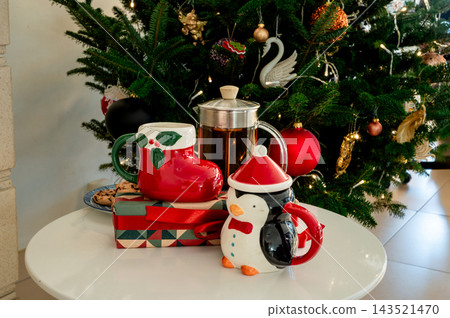 Boot and Penguin Mugs with Gift and French Press by Tree. A red Santa boot mug and a cute penguin mug beside a geometric gift with a red bow and a glass French press of tea before a glowing Christmas Boot and Penguin Mugs with Gift and French Press by Tree. A red Santa boot mug and a cute penguin mug beside a geometric gift with a red bow and a glass French press of tea before a glowing Christmas 143521470