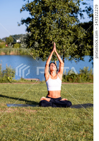 Woman in Seated Pose With Arms Overhead on a Yoga Mat Outdoors. A young Hispanic woman practises a seated pose with arms overhead on the grass beside the water in the evening light, connecting body an 143522229