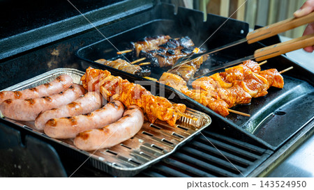 Assorted Meat Skewers and Sausages Cooking on Summer Grill. Sausages in a foil tray beside assorted marinated meat skewers cooking on an outdoor gas barbecue with tongs at a forest cabin. A summer coo Assorted Meat Skewers and Sausages Cooking on Summer Grill. Sausages in a foil tray beside assorted marinated meat skewers cooking on an outdoor gas barbecue with tongs at a forest cabin. A summer coo 143524950