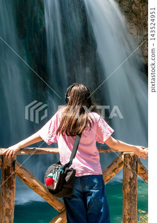 Teen Girl Looking at a Waterfall in the Park. A teenage girl in a pink t shirt stands at a wooden railing looking at a waterfall in the Monasterio de Piedra natural park in Aragon, Spain, a traveler e 143524951