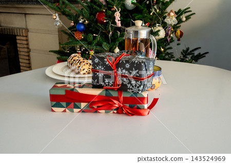 Two Stacked Christmas Gifts with French Press by Glowing Tree. Two festive gifts, a black snowflake box and a geometric present with a red bow, stacked beside a glass French press of tea Two Stacked Christmas Gifts with French Press by Glowing Tree. Two festive gifts, a black snowflake box and a geometric present with a red bow, stacked beside a glass French press of tea 143524969